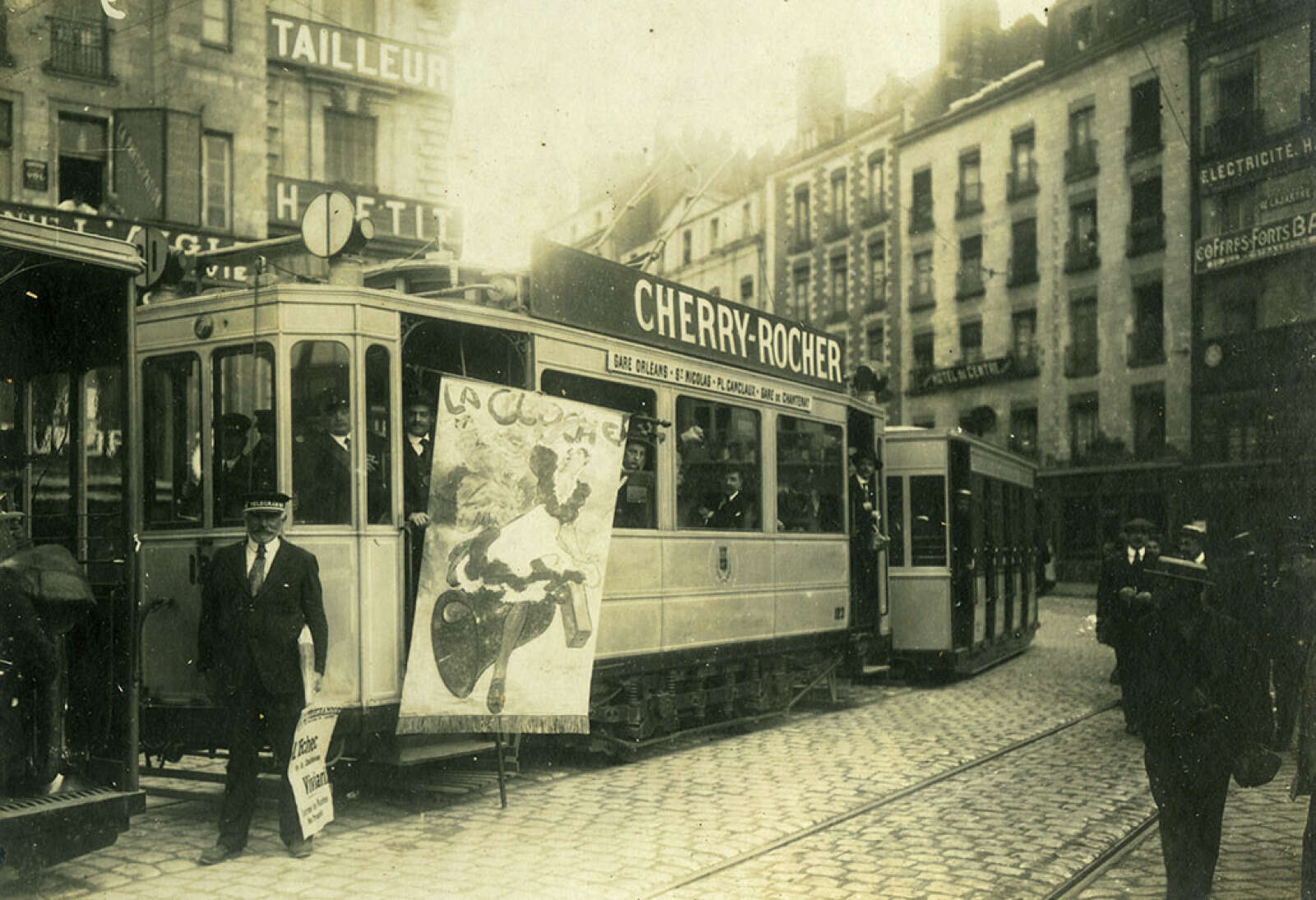 Vue des sociétaires de La Cloche déployant la bannière de l'association à bord d'un tramway, place du Commerce, au départ d'une excursion aux Sables-d'Olonne pour y tenir leur banquet annuel.